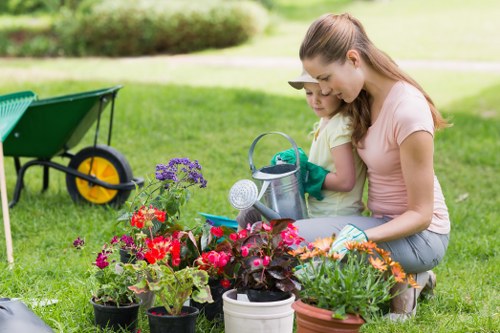 A woman and a young girl are kneeling on a well-maintained, lush green lawn in a garden, surrounded by an array of vibrant flowering plants in pots, including red, purple, and orange blooms. They are engaged in gardening activities, with the woman holding a small watering can and the girl wearing gardening gloves, indicating they are watering or tending to the plants. In the background, there is a garden wheelbarrow with bright yellow wheels, partially visible on the left side, and a neatly clipped hedge lining the edge of the garden space. The environment appears bright and sunny, with natural daylight illuminating the scene, suggesting a pleasant day suitable for outdoor gardening in the Vauxhall area. The garden includes features like grass, flower beds, and garden tools, reflecting well-maintained outdoor space typical of residential gardens in London Borough of Vauxhall, with a focus on gardening and lawn care services such as those offered by Lawn Mowing Vauxhall.